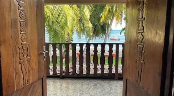 A wooden door with intricate carvings, leading to a balcony with a view of palm trees and the sea.