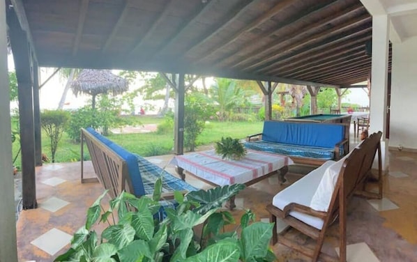 A covered outdoor seating area with wooden furniture and a view of a garden.