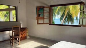 A room with a bed, a desk, and a chair. There is a window with a view of palm trees and the sea. - Villa Carla - Grandiose stays by the sea (Antsiranana)