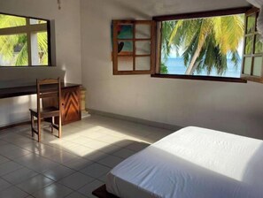 A room with a bed, a desk, and a chair. There is a window with a view of palm trees and the sea.