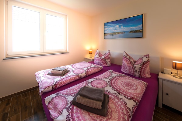 A bedroom with two beds, a window, a nightstand with a lamp, and a painting on the wall.