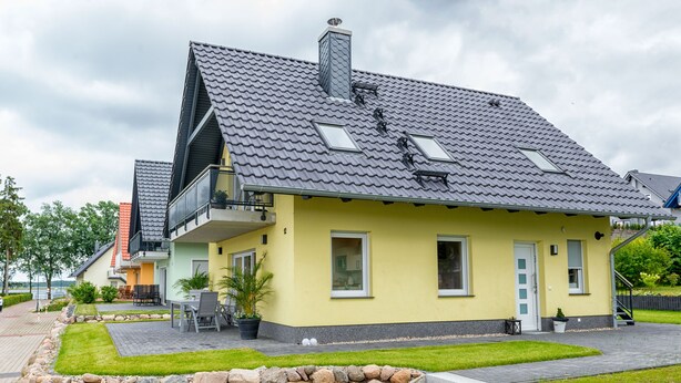 A yellow house with a grey tiled roof, a balcony, and a stone wall in the front yard.