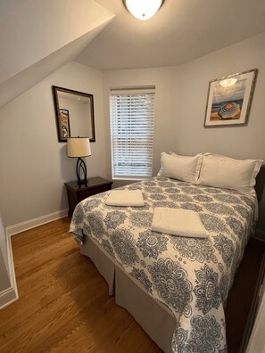 Un dormitorio con cama, mesita de noche, lámpara, ventana con persianas, un cuadro y suelo de madera.