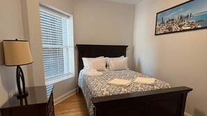 Une chambre à coucher avec un lit, une table de chevet, une lampe, une fenêtre avec des stores et un tableau représentant un paysage urbain accroché au mur.