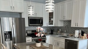 Une cuisine moderne avec des meubles blancs, des appareils en acier inoxydable, un îlot central doté d’un plan de travail en verre et deux luminaires suspendus.