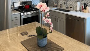 A modern kitchen with grey cabinets, stainless steel appliances, a central island with a vase of pink orchids, and pendant lighting. - Wrigleyville Inn - Unit 3 (Chicago)
