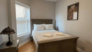 A bedroom with a bed, bedside table, lamp, window, and artwork on the wall.