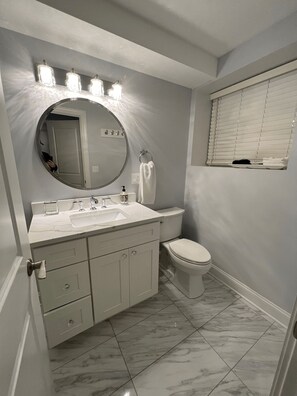 Une salle de bain avec un meuble-lavabo blanc, un miroir rond, des toilettes et une fenêtre avec des stores.