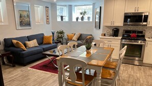 Un salon moderne avec un canapé bleu foncé, des chaises blanches et une table à manger en bois. On y trouve également un coin cuisine doté d’appareils électroménagers en acier inoxydable et d’une bouilloire rouge posée sur la cuisinière.
