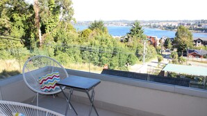 A balcony with a white wicker chair and a small table, overlooking a scenic view of a lake and distant buildings.