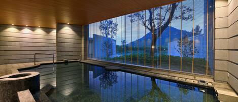 A modern indoor pool with a clear view of the outdoors through a large window, a wooden ceiling, and a stone wall.