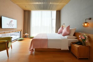 Ein Hotelzimmer mit Bett, Sessel, Nachttisch, Fernseher und einem Fenster mit Vorhängen.