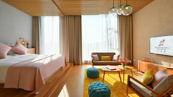 Habitación de hotel con cama, sofá, mesa de centro, otomanes y una pantalla de televisión mostrando el nombre del hotel.