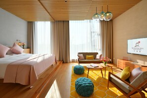 Habitación de hotel con cama, sofá, mesa de centro, otomanes y una pantalla de televisión mostrando el nombre del hotel.