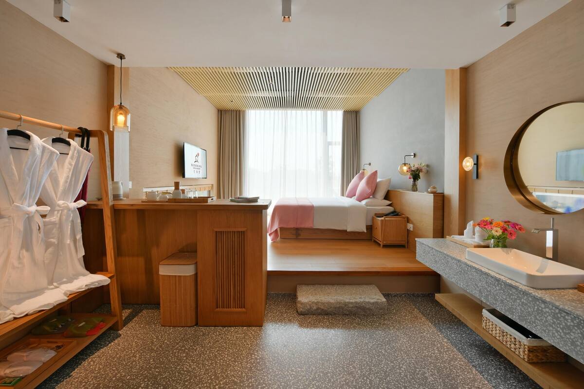A modern hotel room with a wooden desk, a bed with pink bedding, a large mirror, and a bathroom with a granite countertop.