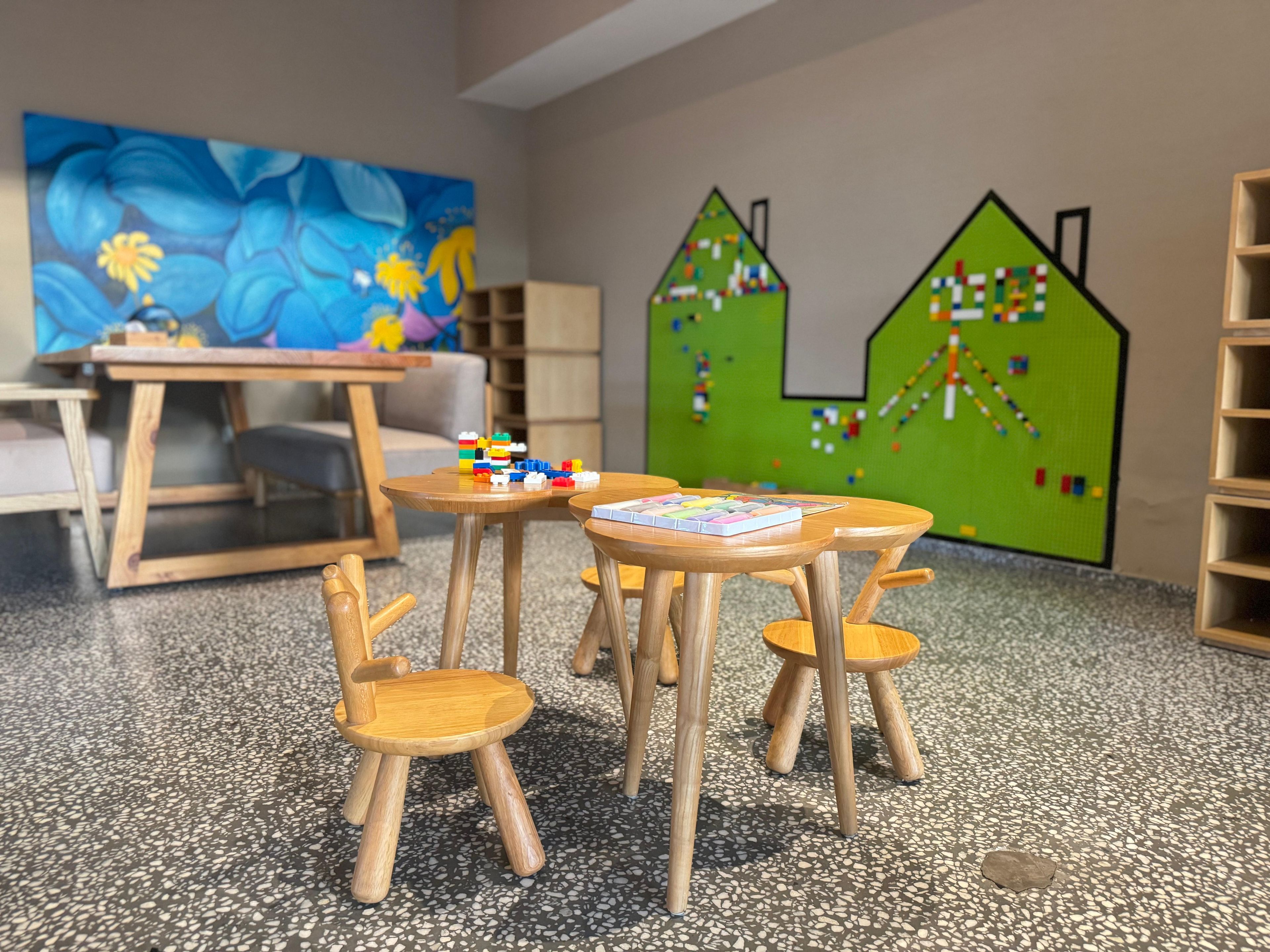 Un salón de juegos infantiles con mesas y sillas de madera, un cuadro colorido y una pared verde con un rompecabezas con forma de casa.