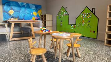 Un salón de juegos infantiles con mesas y sillas de madera, un cuadro colorido y una pared verde con un rompecabezas con forma de casa.