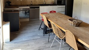 Et moderne køkken med træbjælker, et langt spisebord, fletstole og et sort køleskab.