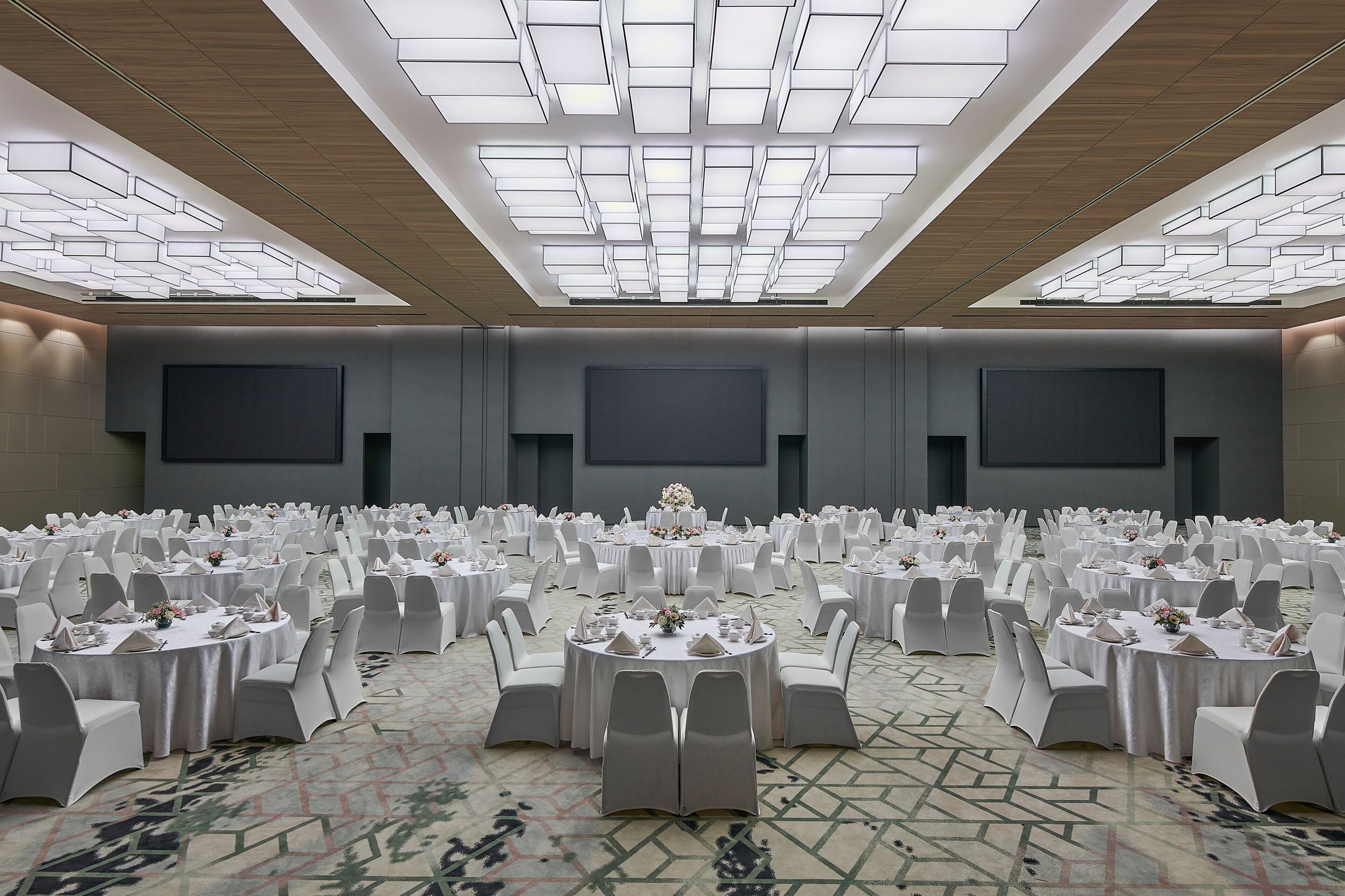 Un amplio salón de conferencias con mesas redondas y sillas blancas, alfombra estampada y un diseño moderno del techo.