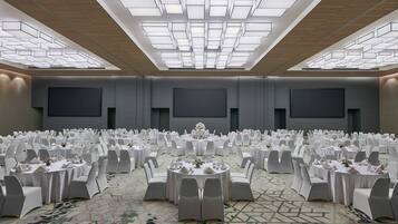Un amplio salón de conferencias con mesas redondas y sillas blancas, alfombra estampada y un diseño moderno del techo.