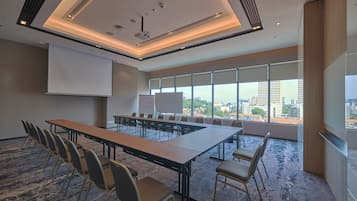 Un salón de conferencias con una larga mesa, sillas, una pantalla de proyector y grandes ventanas que ofrecen vistas a la ciudad.