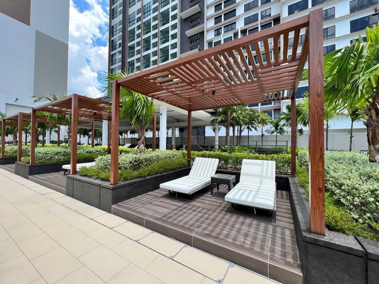 A modern outdoor lounge area with wooden pergolas, white lounge chairs, and low-rise buildings in the background.