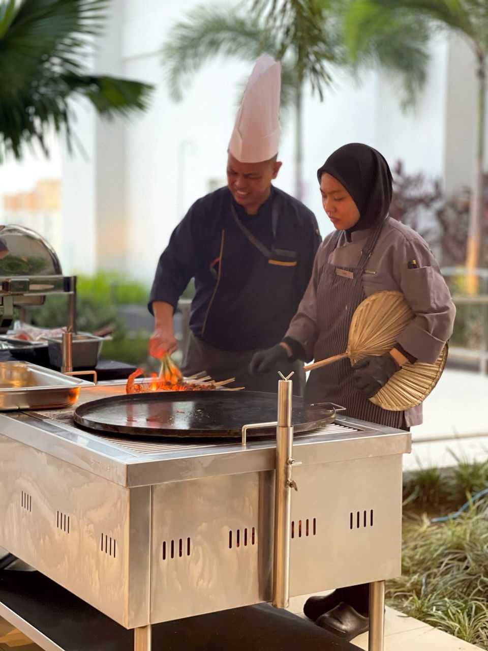 Un chef preparando comida en una parrilla grande al aire libre, con un hombre ayudando.