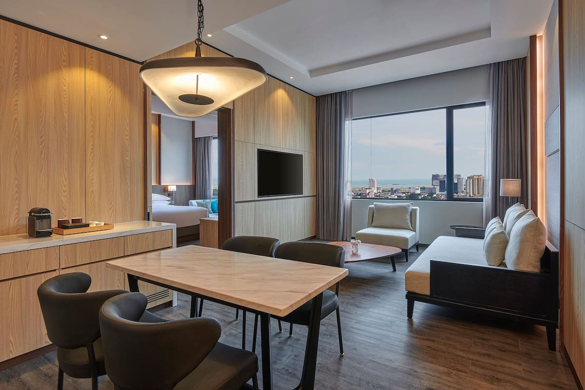 Une chambre d’hôtel moderne dotée d’un coin repas, d’un canapé, d’un téléviseur fixé au mur et d’une grande fenêtre donnant sur la ville.