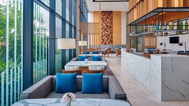 Un hall d’hôtel moderne, doté d’espaces aménagés avec des sièges, d’un comptoir de réception et d’une grande fenêtre offrant une vue sur la verdure.