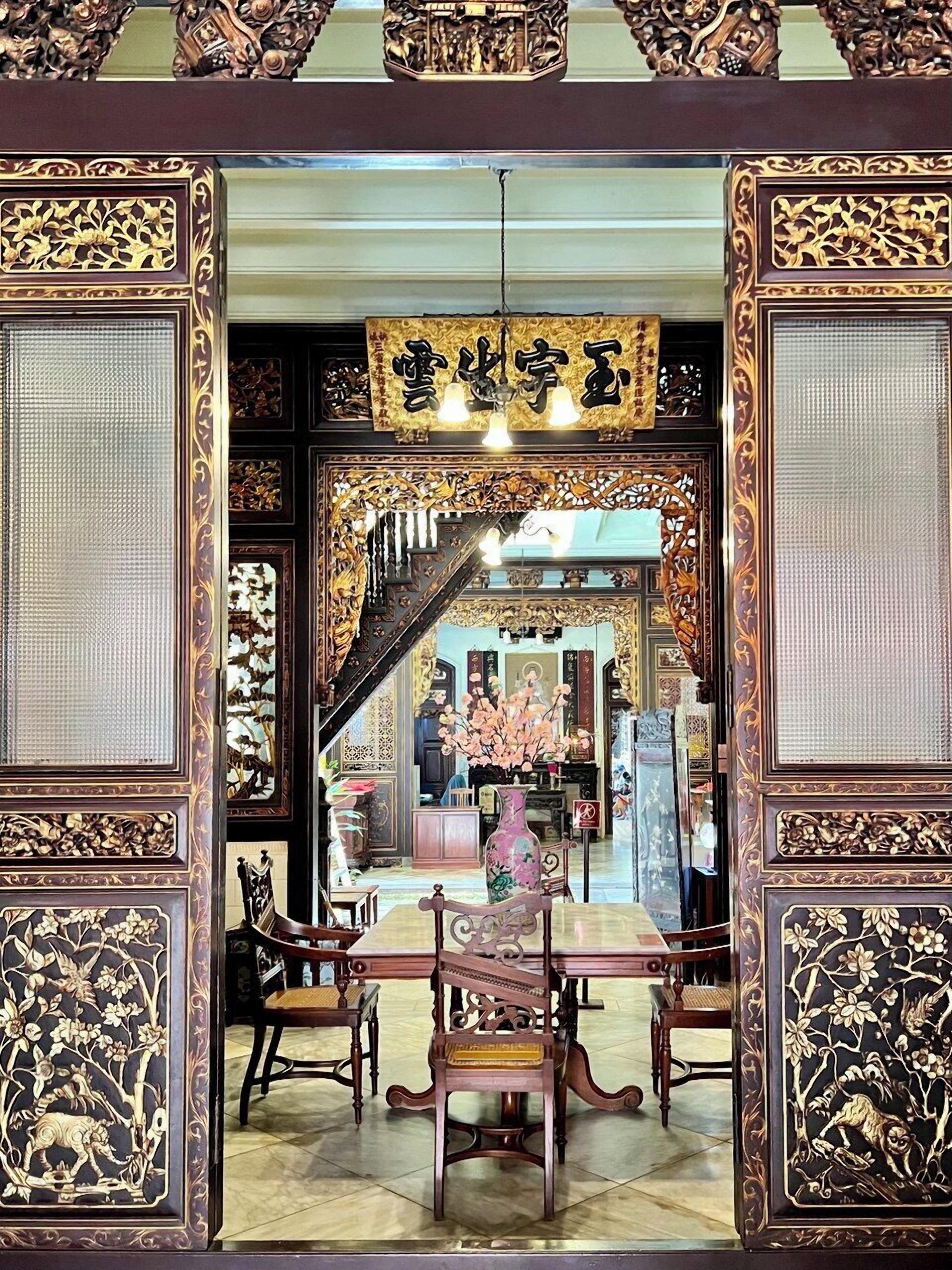 Interior tradicional chinês com elaboradas esculturas em madeira, uma mesa central com cadeiras e uma escadaria que leva a um andar superior.