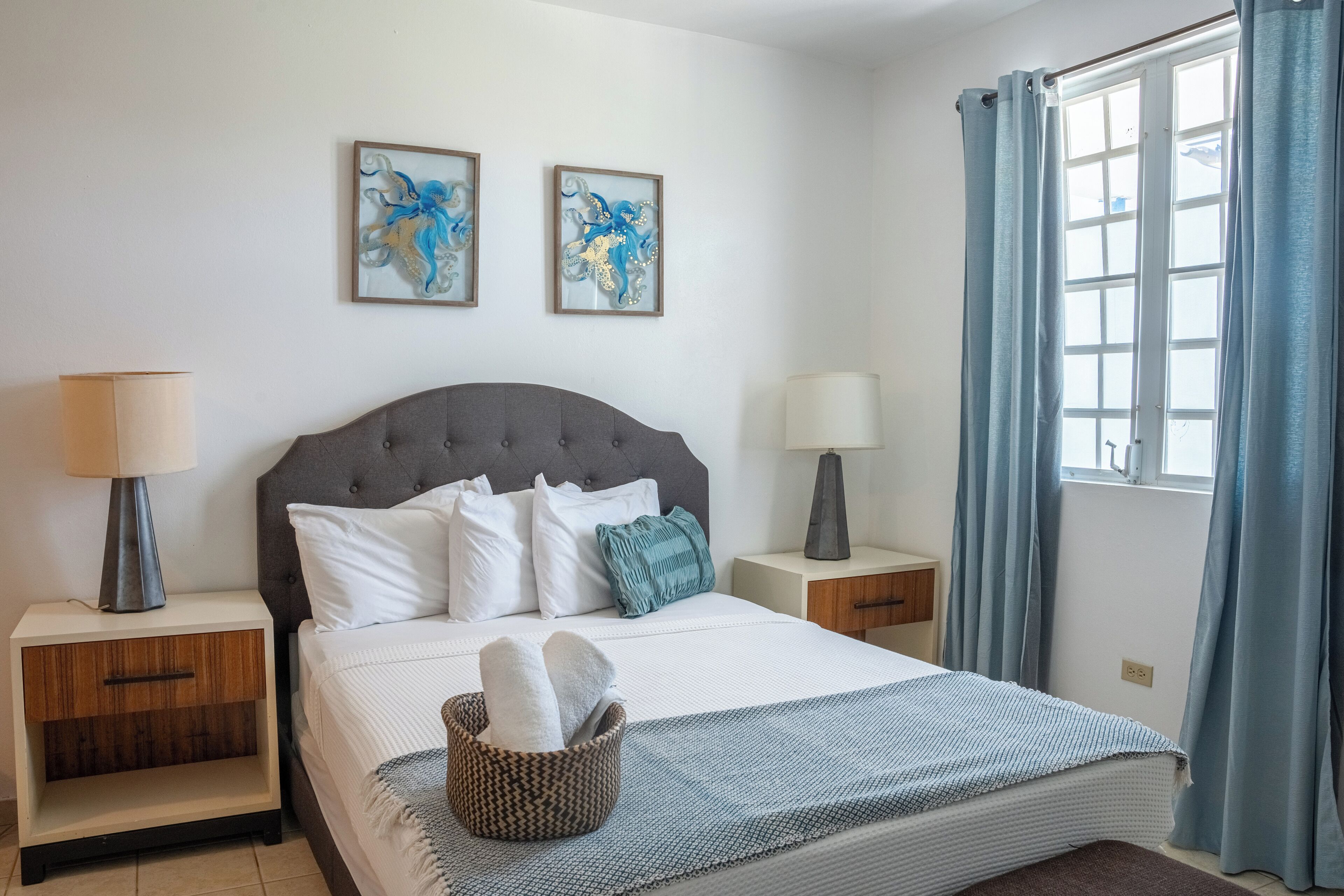 Een netjes ingerichte slaapkamer met een groot bed, twee nachtkastjes, een raam met gordijnen en twee ingelijste kunstwerken aan de muur.