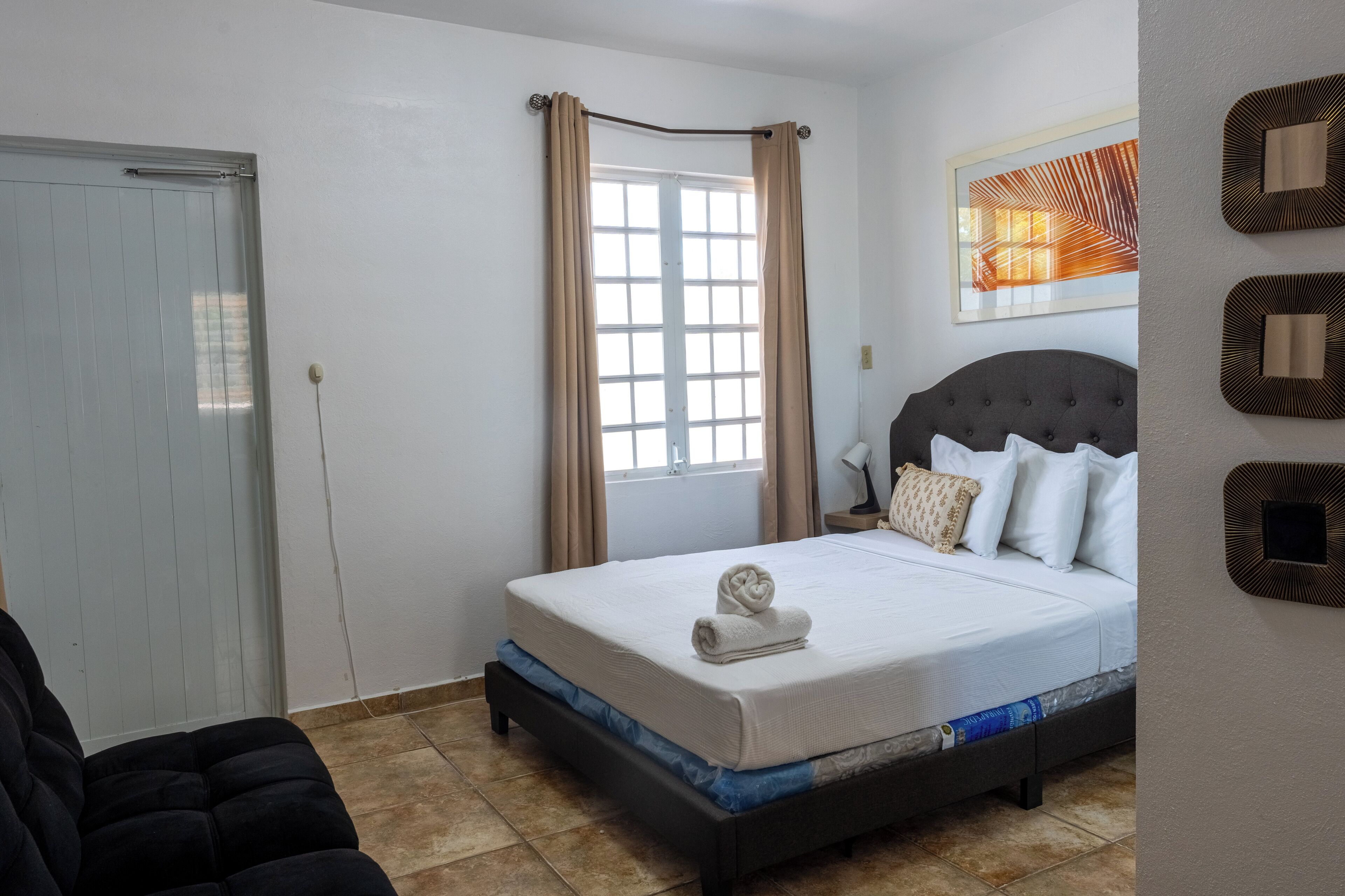 A bedroom with a bed, a nightstand, a window with curtains, a door, and a wall with a framed picture.