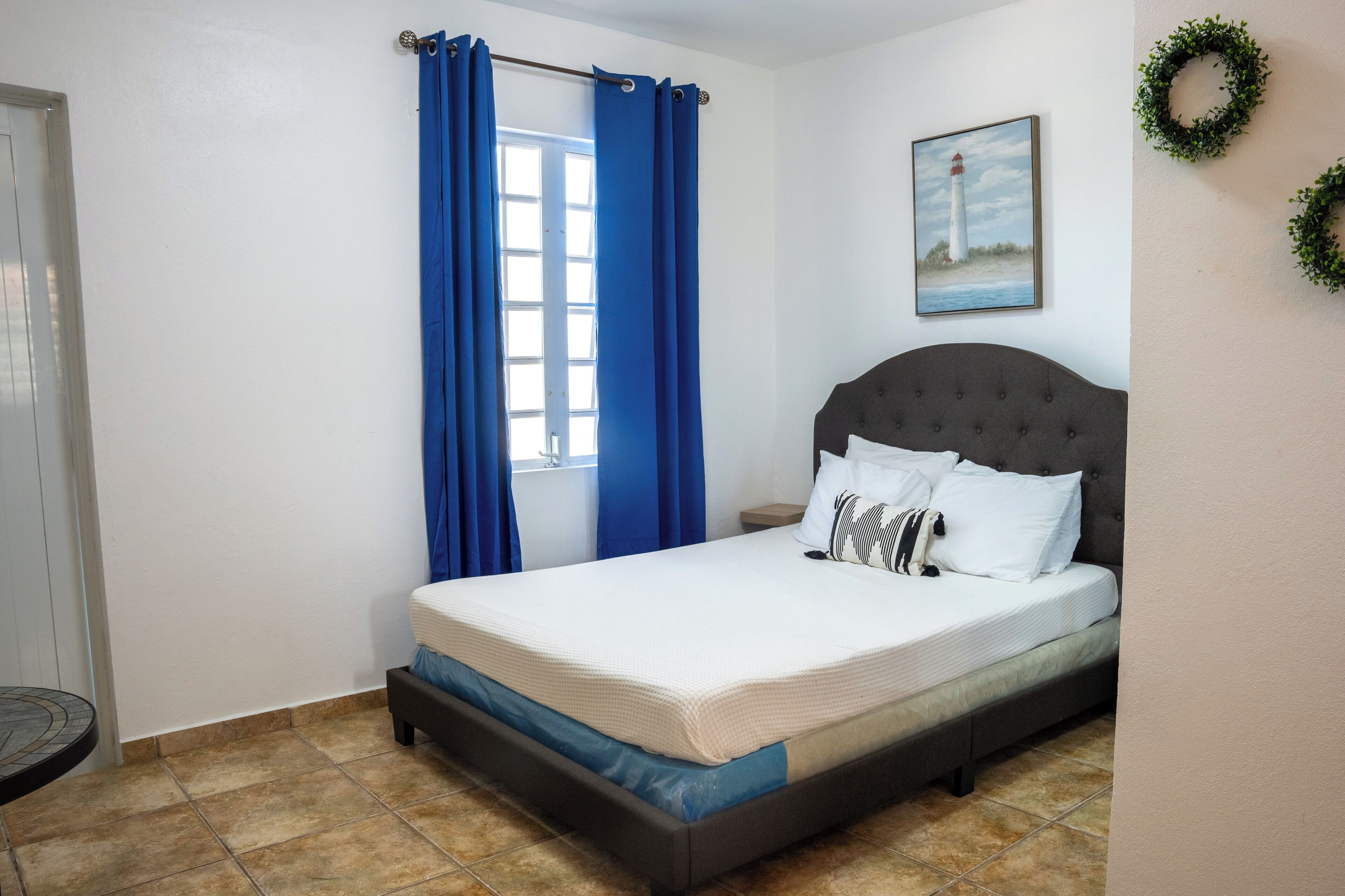 A bedroom with a bed, blue curtains, a headboard, a painting of a lighthouse, and a wall-mounted wreath.