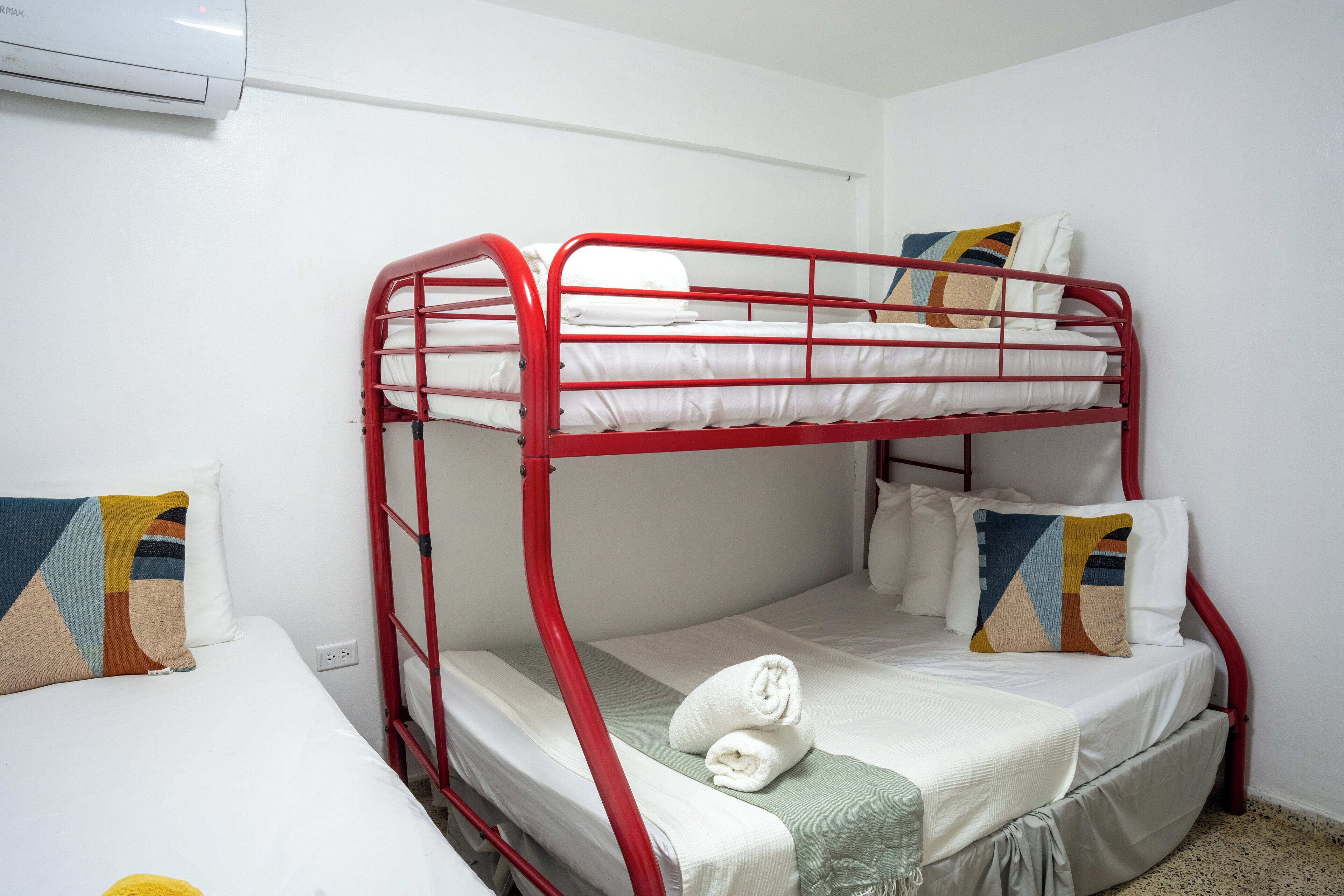 A bunk bed with a red frame, white bedding, and a green blanket. There is a white pillow with a geometric pattern on the lower bunk.