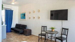 Ein kompakter Wohnbereich mit Sofa, kleinem Tisch, zwei Stühlen, einem an der Wand befestigten Fernseher und einem Deckenventilator.