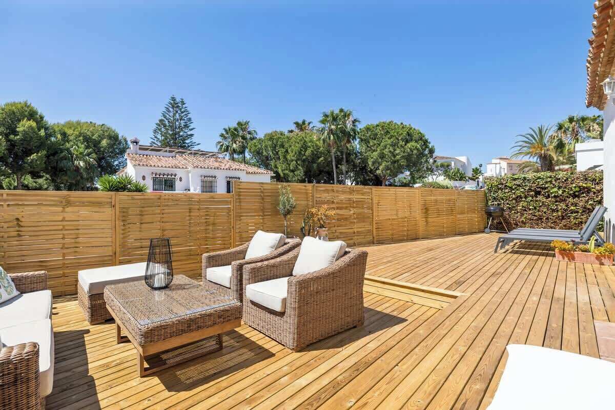 Una terraza de madera con mobiliario de mimbre, un edificio blanco al fondo y una valla de madera.