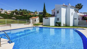 Ein Swimmingpool mit glasklar blauem Wasser, umgeben von einem weißen Gebäude mit einem Ziegeldach und einem gepflegten Rasen mit einer Einfriedung.