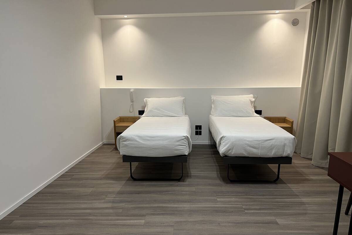 Habitación de hotel moderna con dos camas individuales, una mesita de noche de madera, una mesilla auxiliar y un teléfono en la pared.