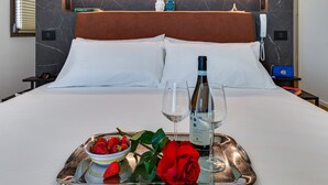 Un lit parfaitement fait, avec du linge de lit blanc, un plateau sur lequel se trouvent une bouteille de vin, deux verres, un bol de fraises et une rose rouge, dans une chambre d’hôtel.