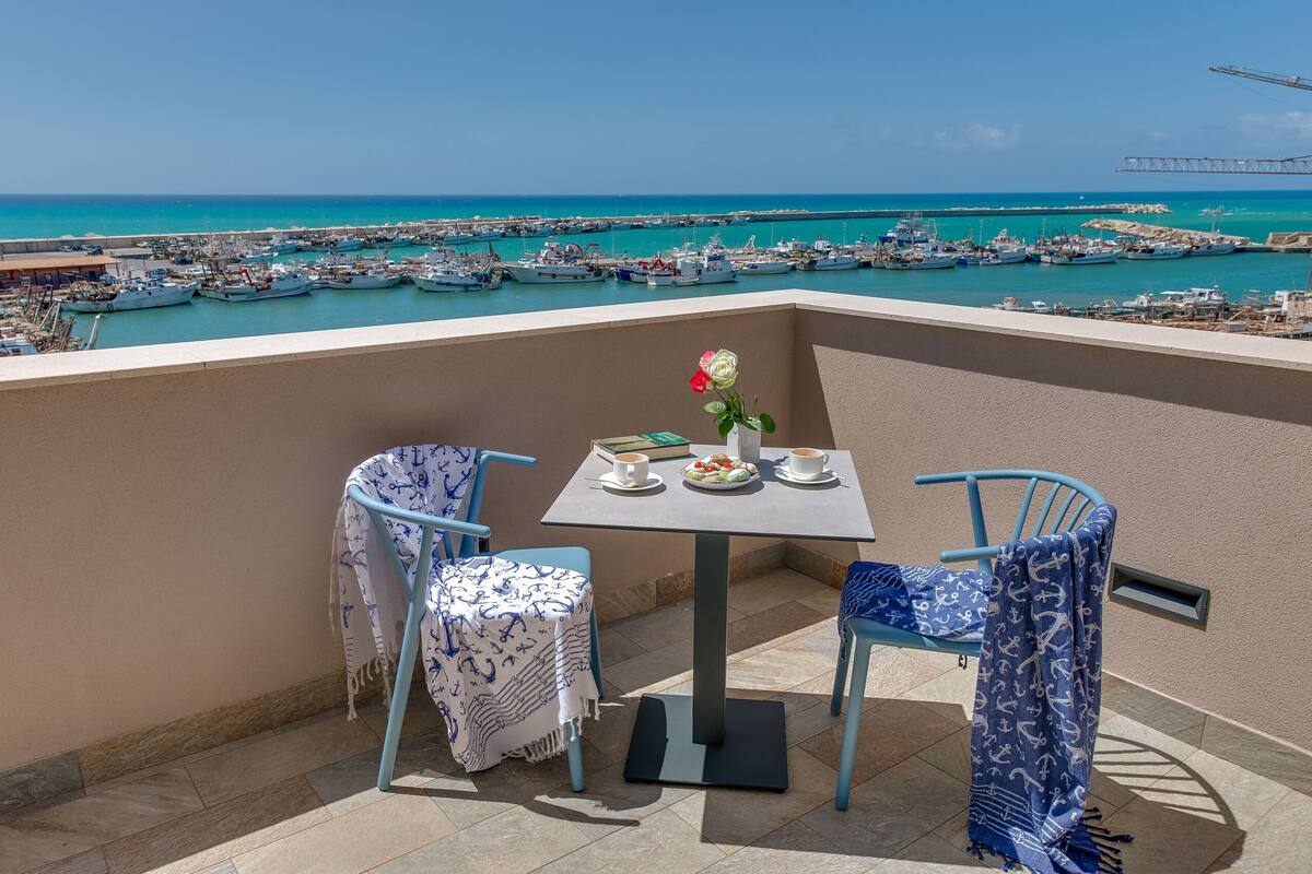 Un balcón con dos sillas, una mesa y una flor. El balcón tiene vistas a un puerto deportivo con barcos.
