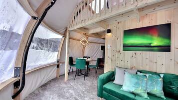 A cozy interior with a green sofa, a dining table with chairs, and a painting of the Northern Lights.