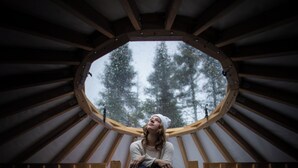 Una persona sentada en una escalera de madera dentro de una habitación circular con una ventana, rodeada de vigas de madera.