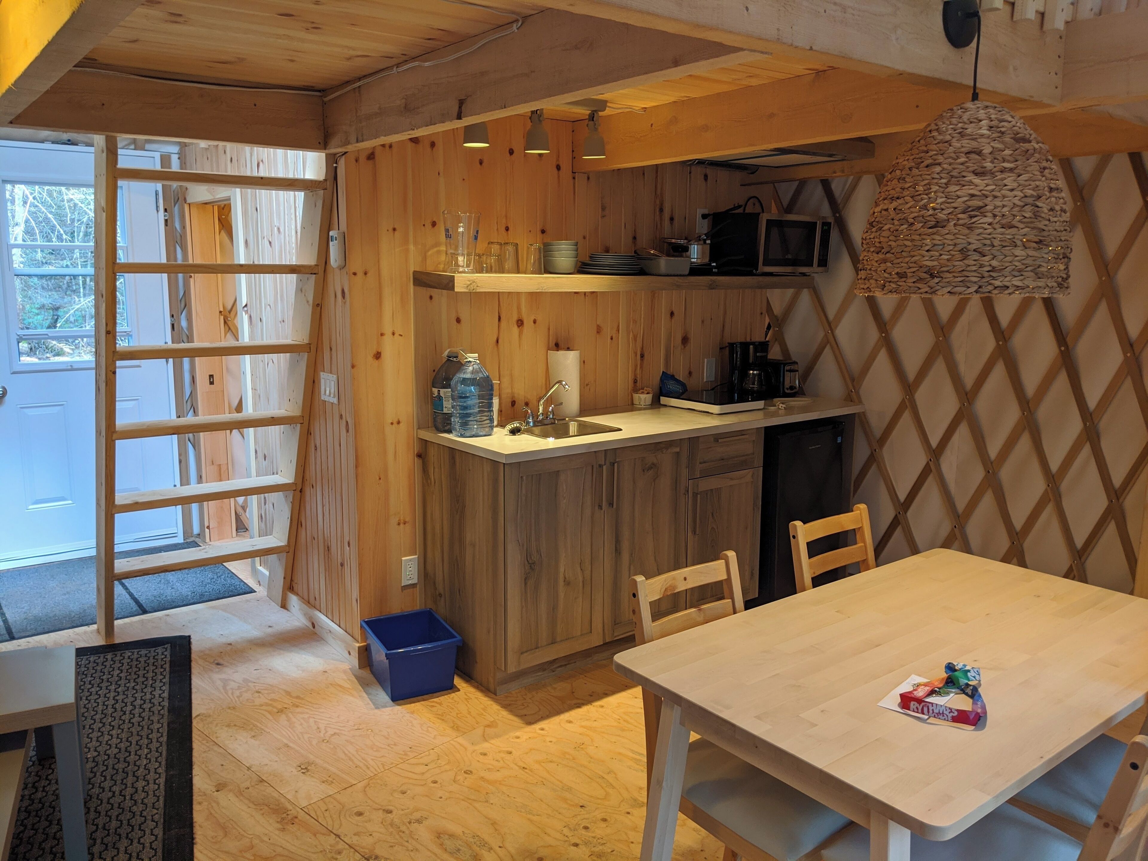 A compact kitchen with wooden cabinets, a sink, and a microwave. There is a dining table with chairs and a blue trash bin.