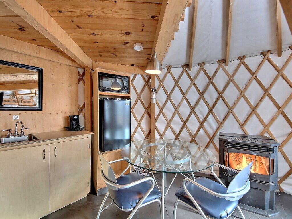 Un intérieur de chalet chaleureux avec un plafond en bois, une table en verre entourée de chaises, un coin cuisine et un poêle à bois.