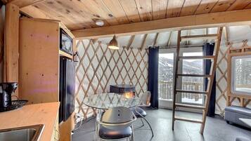 Interior acolhedor de uma cabana, com teto de madeira, área de jantar com mesa e cadeiras, lareira e vista para montanhas cobertas de neve através de grandes janelas.