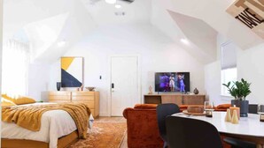 Ein modernes Schlafzimmer im Loft-Stil mit schrägverdachter Decke, einem Bett mit gelber Decke, einem roten Sofa, einem Esstisch mit Stühlen und einem Fernseher im Hintergrund.