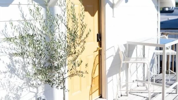 Ein weißes Gebäude mit einer Holztür, einem Balkon mit weißen Stühlen und einem Blumentopf.