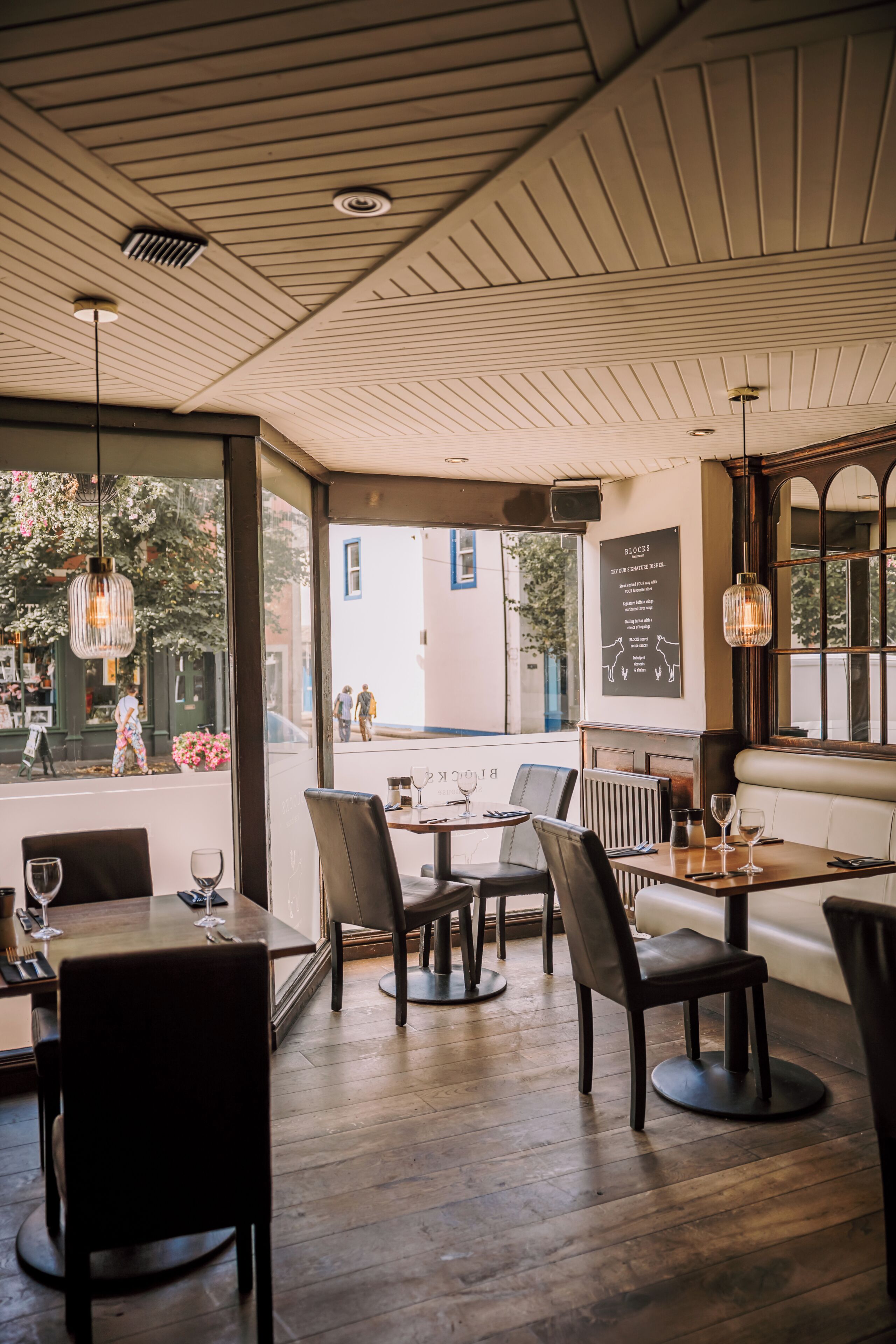 A restaurant interior with wooden floors, tables, chairs, hanging lights, and a view of the street outside.