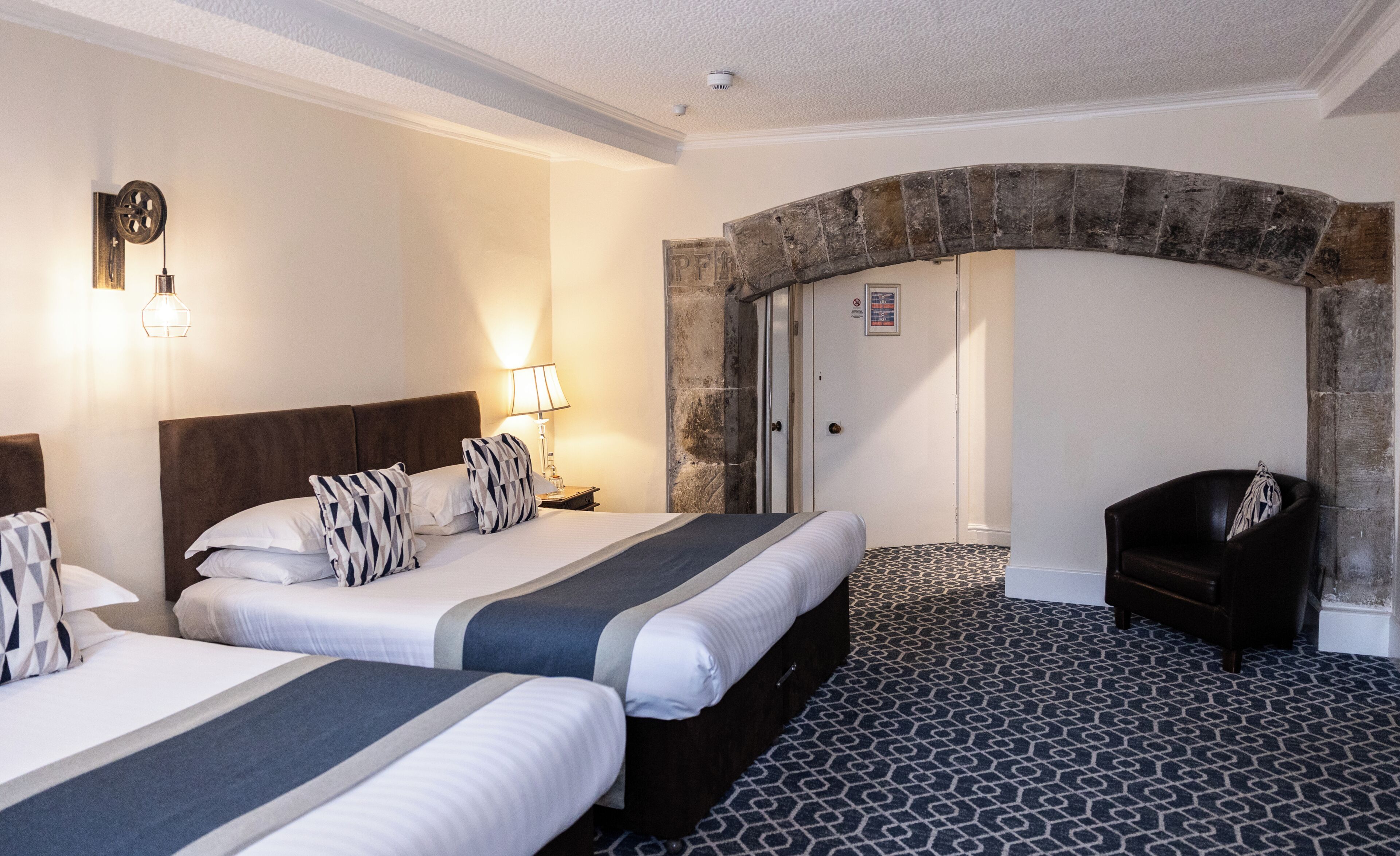 Hotellihuone, jossa on kaksi sänkyä, kivinen holvikaari, tuoli ja kuviollinen matto.