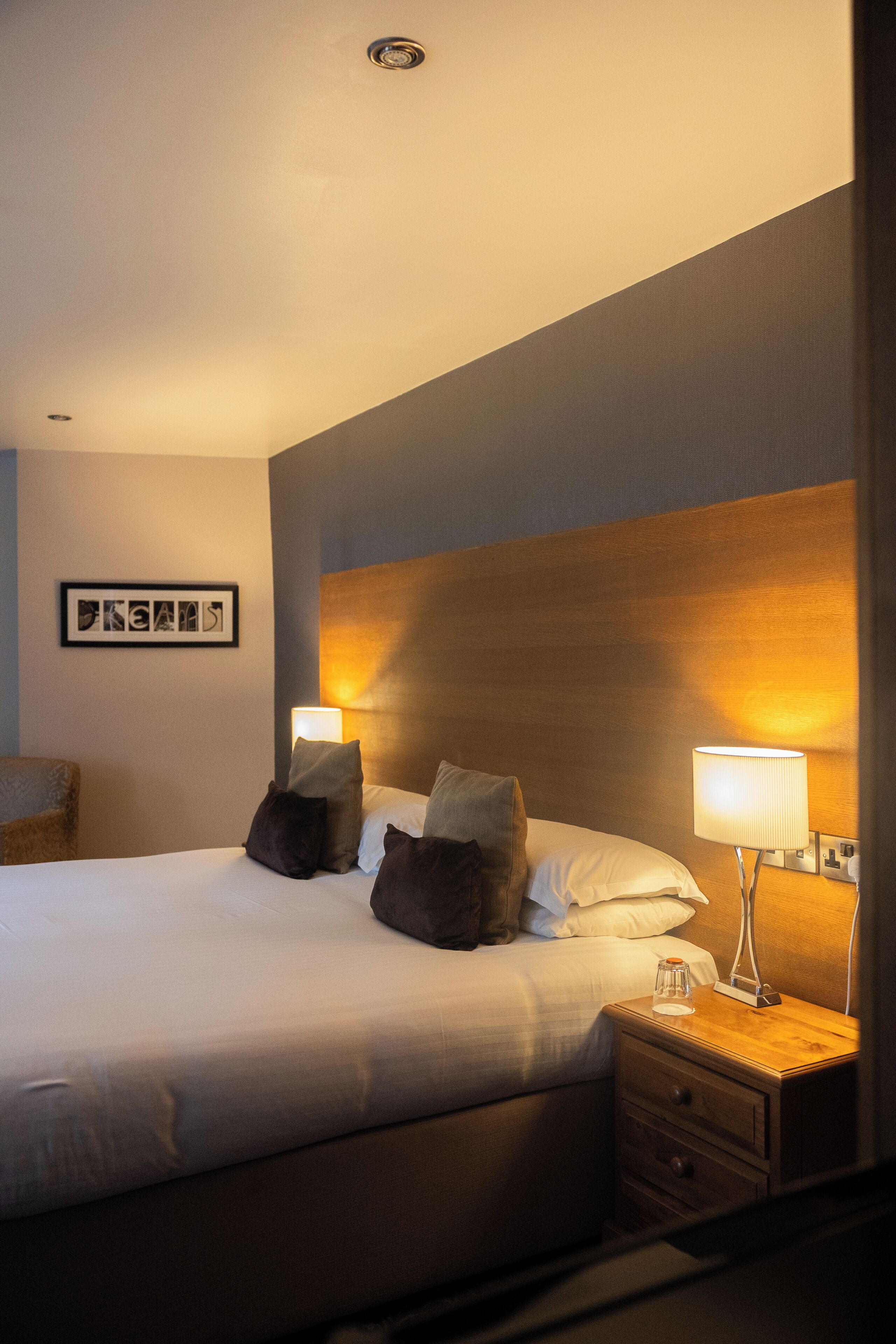Habitación de hotel con una cama grande, dos lámparas de noche, un cabecero de madera, una mesita de noche y un cuadro enmarcado en la pared.